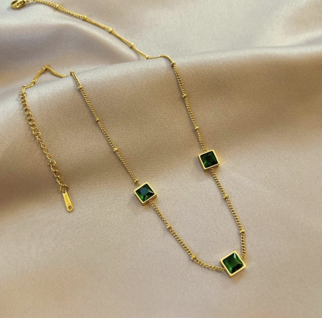 A gold chain necklace with three square green gemstone charms, displayed on a draped beige satin fabric.