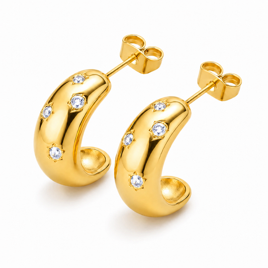 A pair of gold crescent-shaped hoop earrings with small clear gemstones set along the front, displayed on a clean white background.