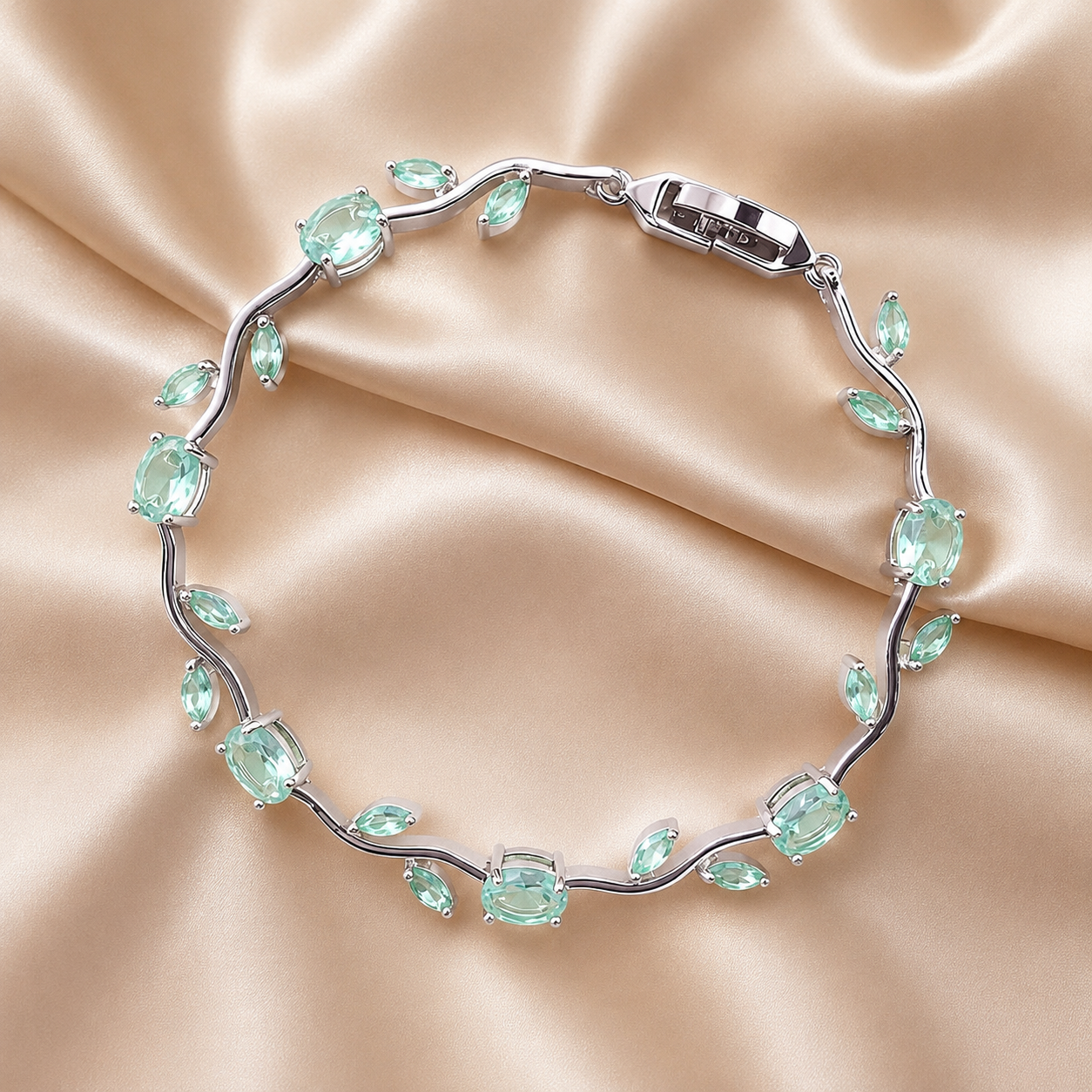 A silver vine-style bracelet with light aqua green marquise and oval crystals, arranged in a circle on a draped beige satin fabric background.Delicate multicolor tulip bracelet in premium gold-plated finish