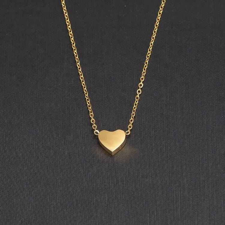 A delicate gold chain necklace with a small, polished heart-shaped pendant, displayed against a dark textured background.